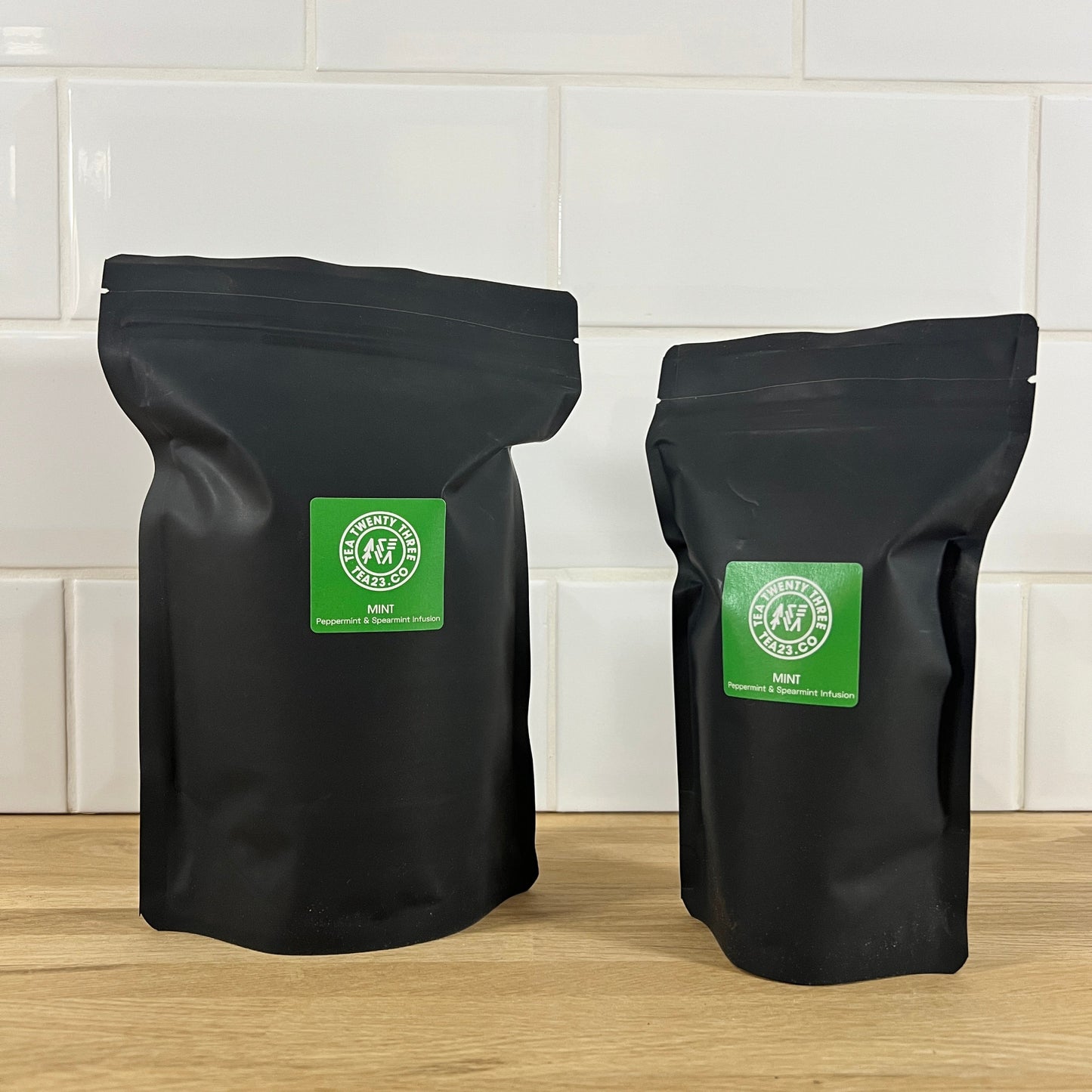 Two black pouches of mint tea with green labels on a wooden surface and tiled wall background