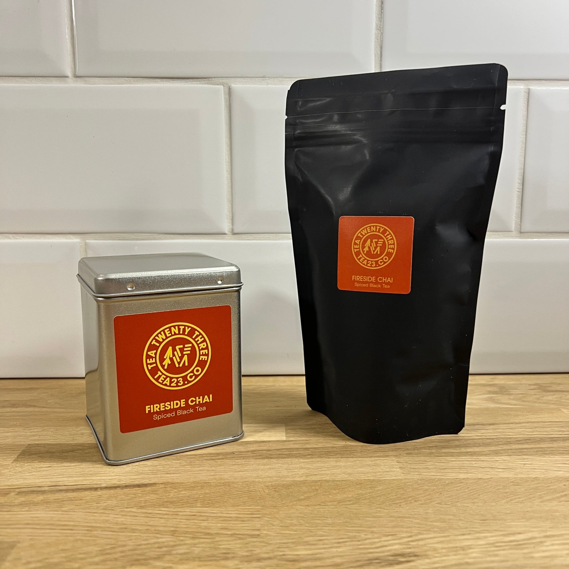 Tea packaging including a black bag and silver tin with a visible brand logo on a wooden surface.