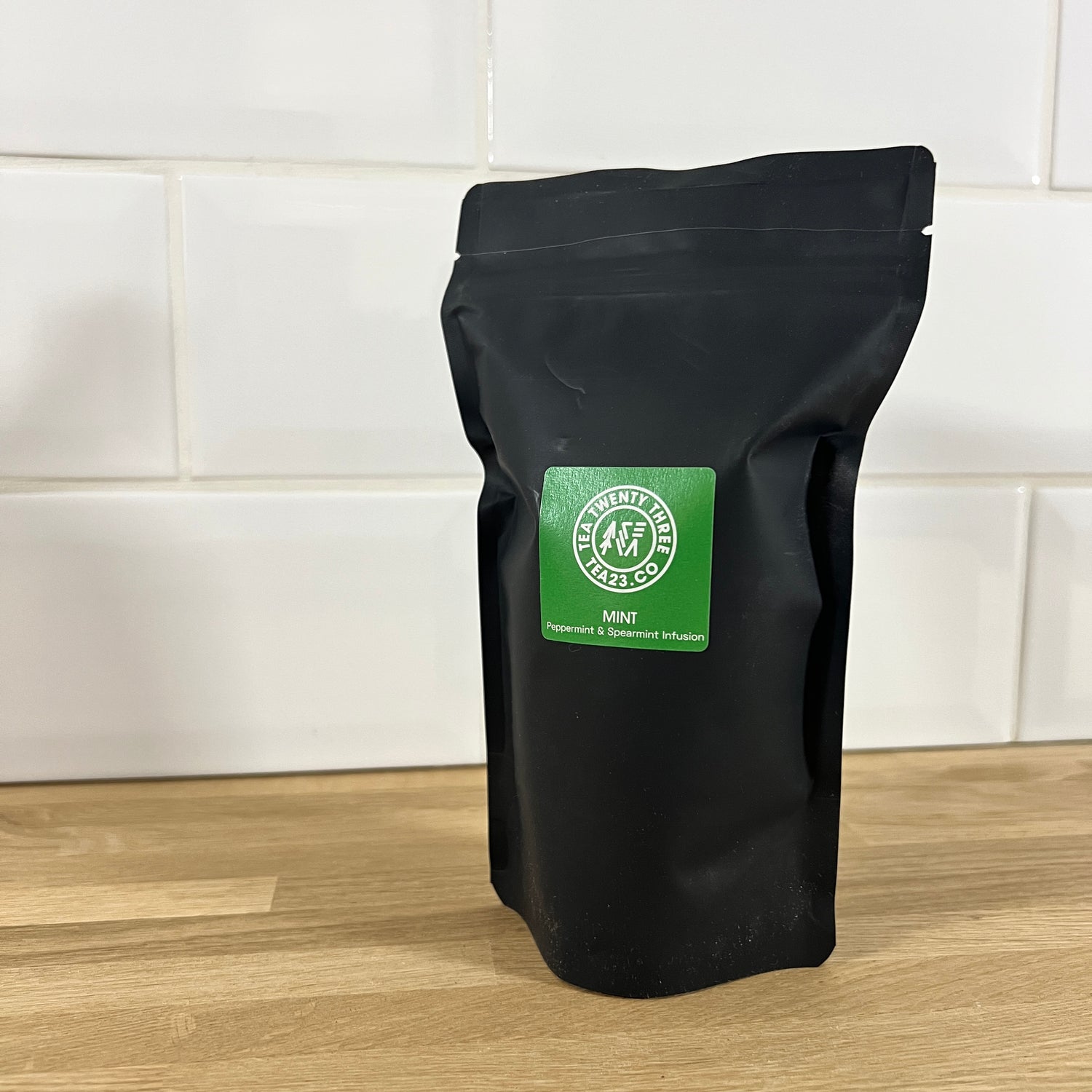 Black pouch of mint tea with a green label on a wooden surface and tiled wall background