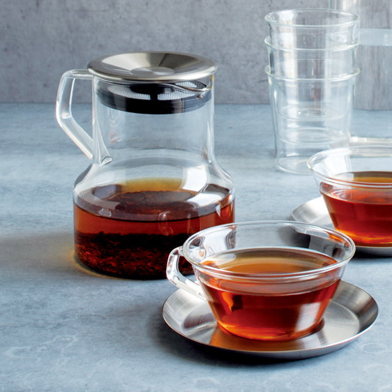 Tea-making set with glass teapot, cups, and saucers on a gray surface.