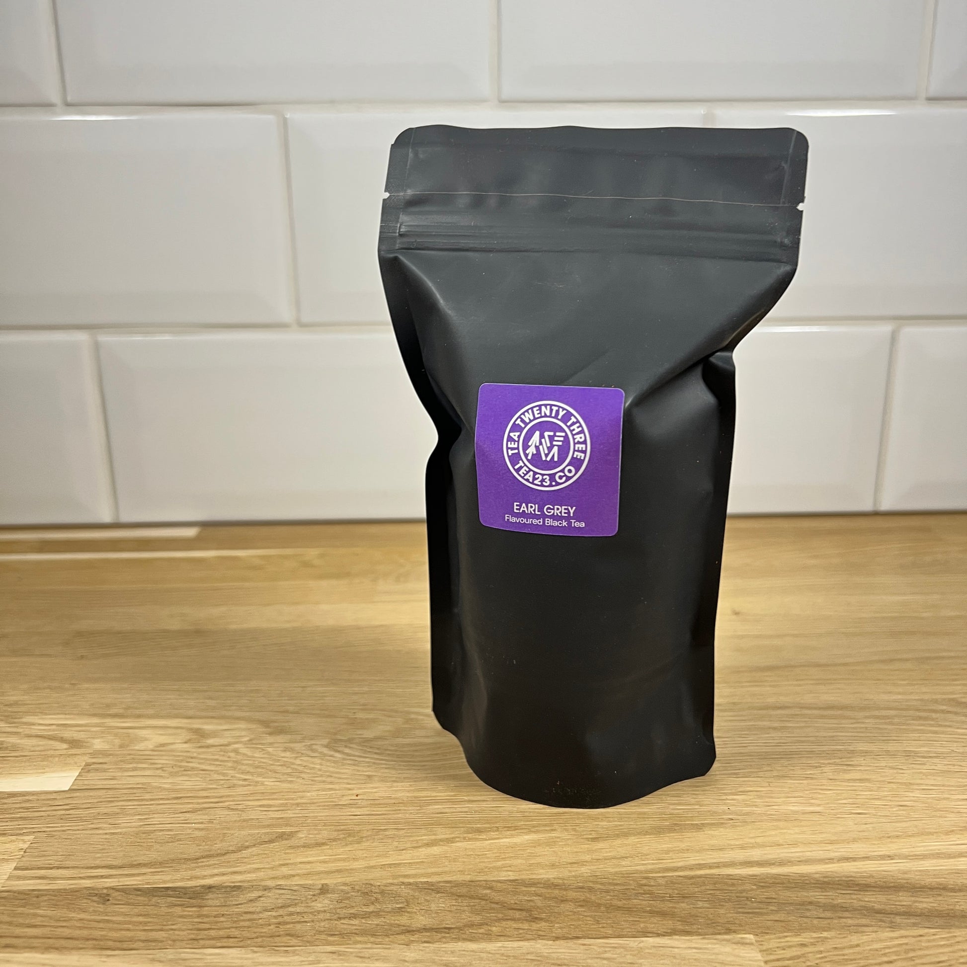 A black pouch of Earl grey tea with a purple label on a wooden surface and tiled wall background