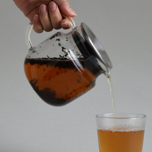 Hand pouring loose leaf tea from a glass teapot into a glass against a neutral background