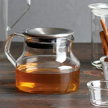 A clear glass teapot filled with amber tea, featuring a stainless steel lid and a black rubber gasket, set on a dark wooden table with other glass containers blurred in the background.