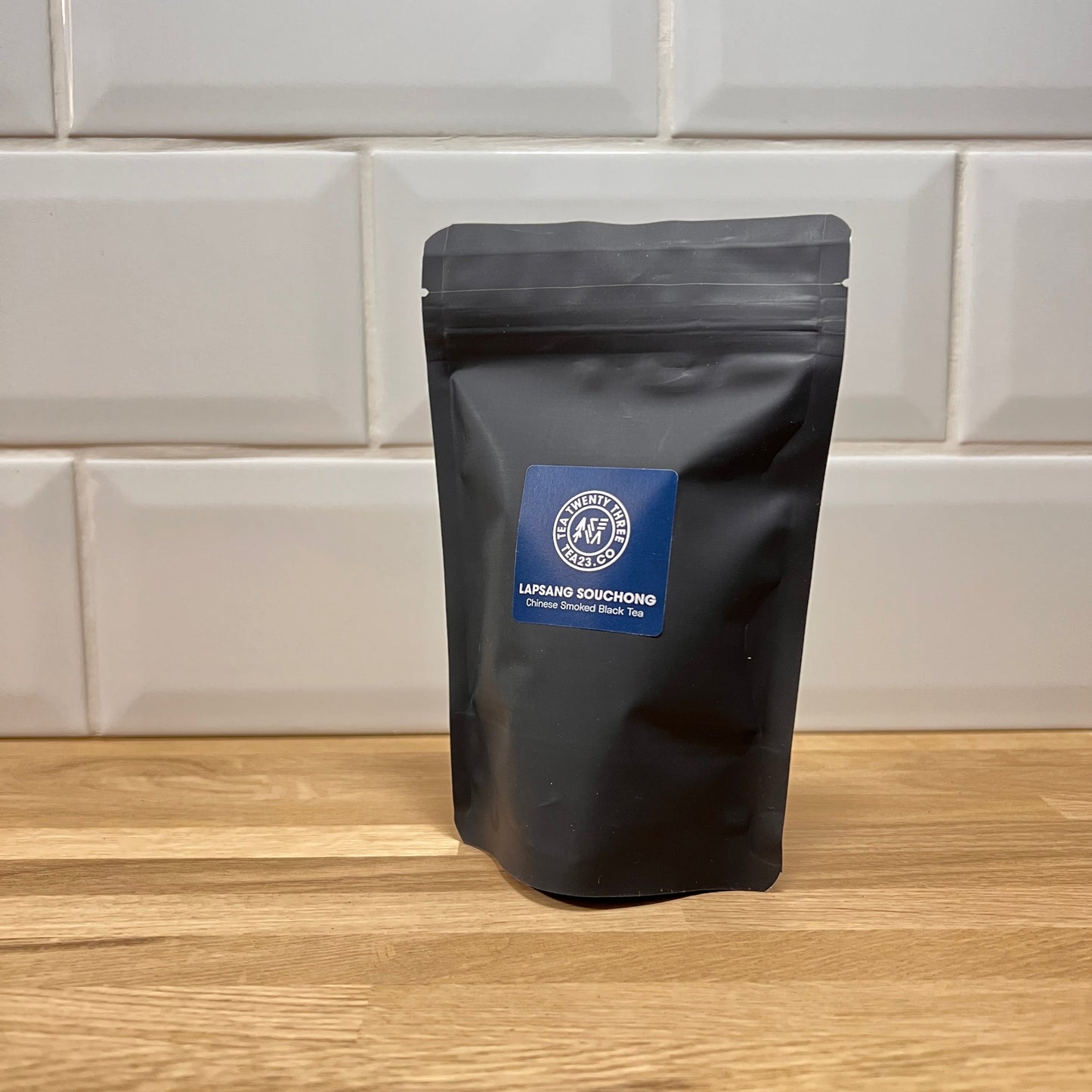 A black pouch of Lapsang Souchong tea with a brand label on a wooden surface and tiled wall background