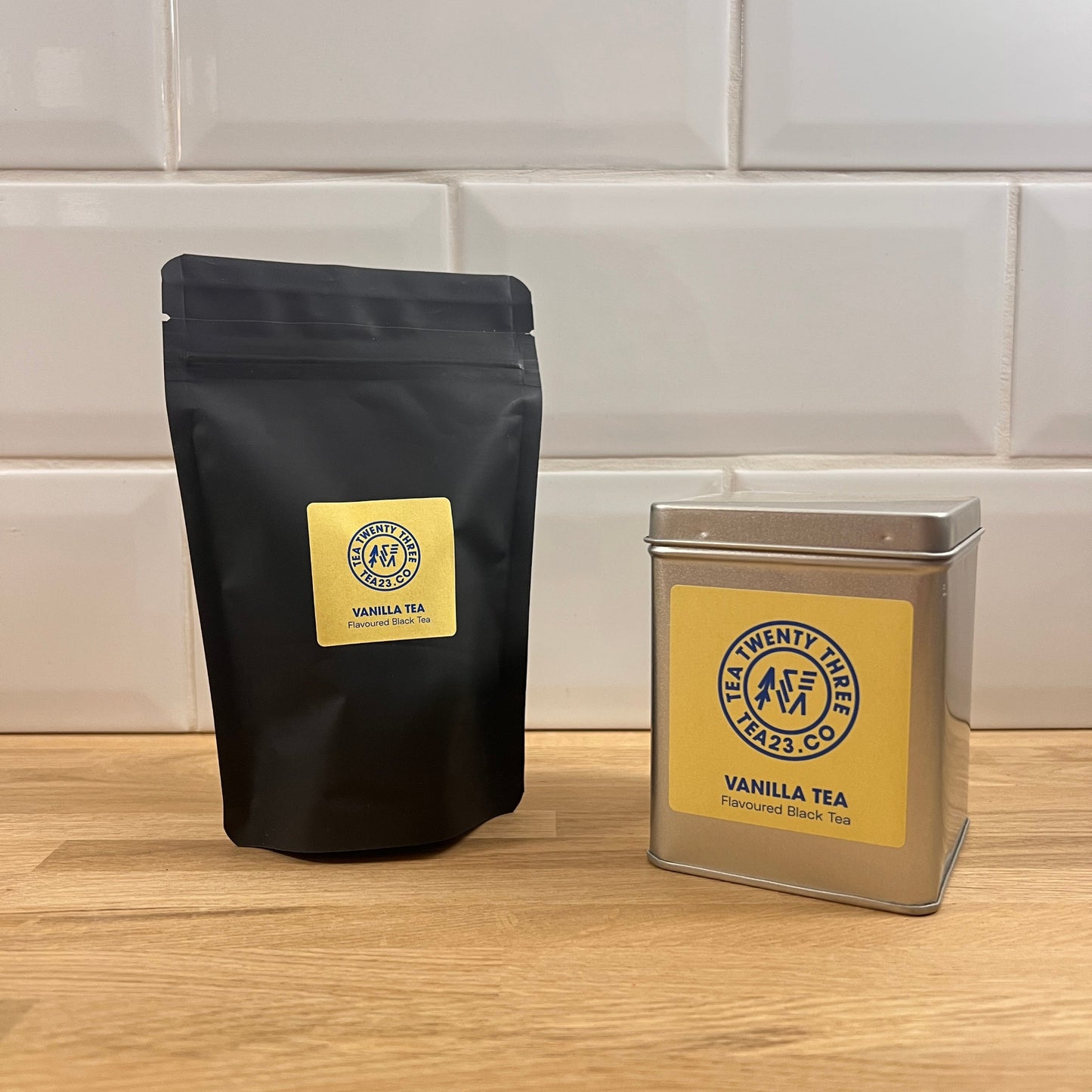 Black tea bag and silver tin of Vanilla Tea from Twenty Three Twenty on a wooden surface with tiled wall background.