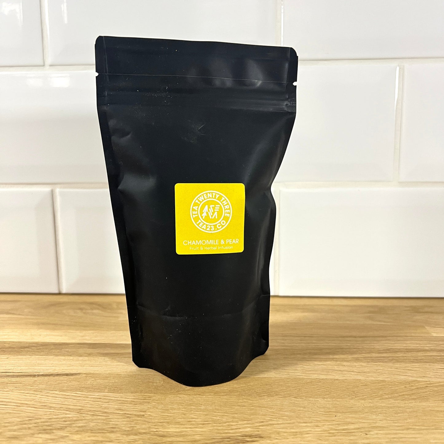A black pouch of chamomile & pear tea with a yellow label on a wooden surface and tiled wall background