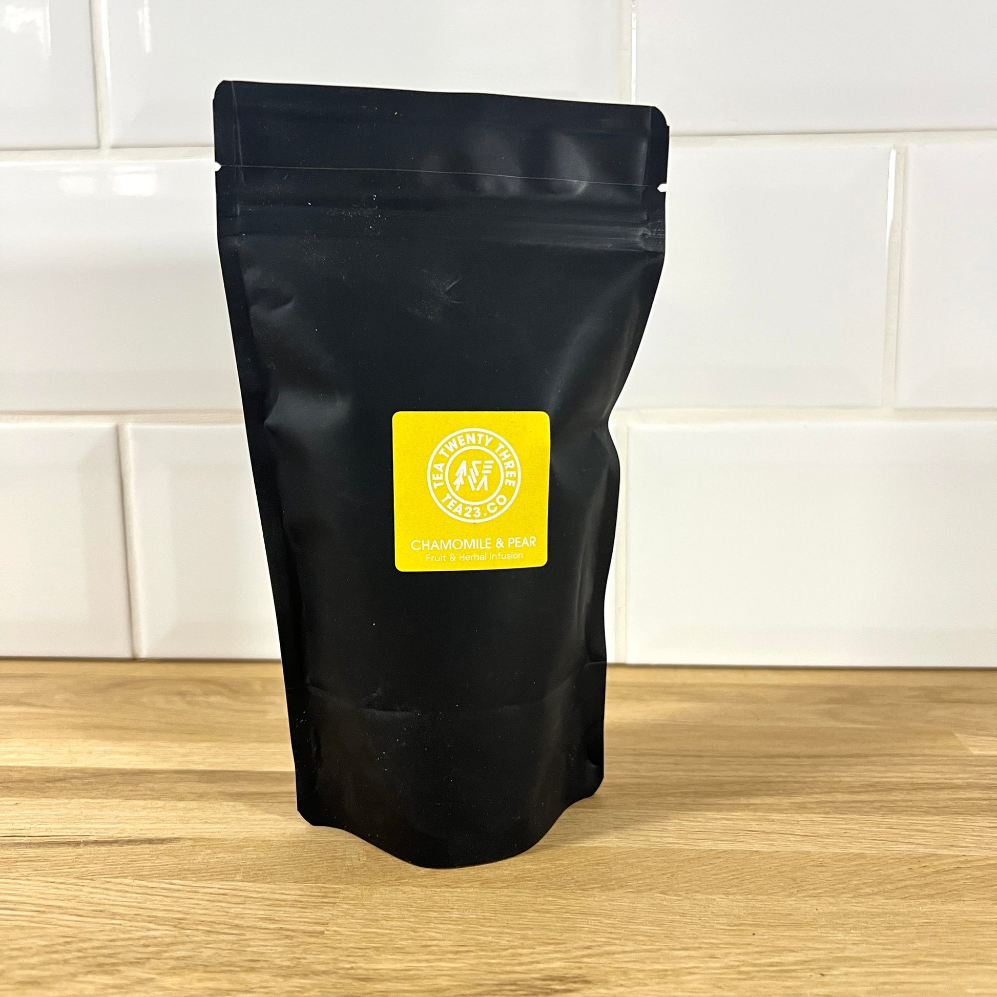 A black pouch of chamomile & pear tea with a yellow label on a wooden surface and tiled wall background