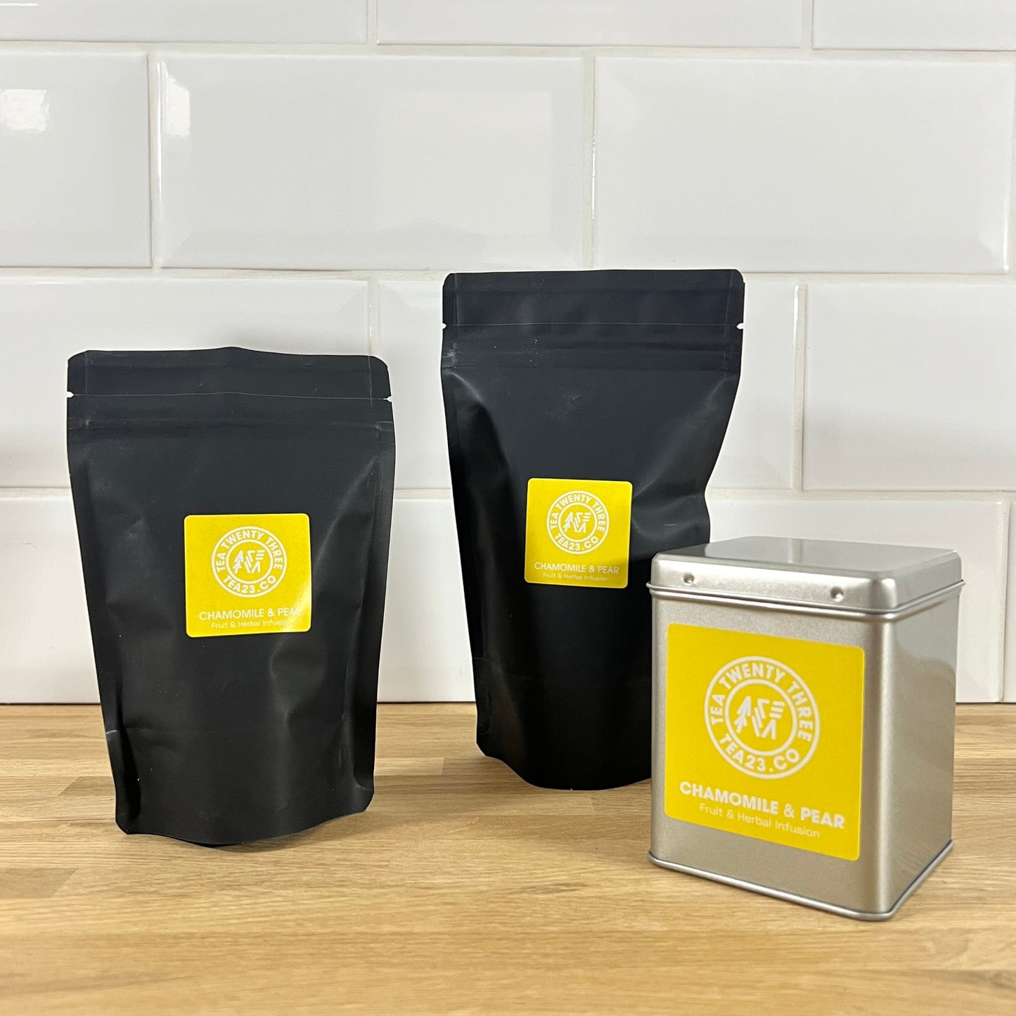 Two black pouches and a silver tin with yellow labels on a wooden surface and tiled wall background.